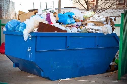 A large blue industrial skip filled with assorted waste materials, including cardboard boxes, plastic packaging, and crumpled paper, situated on a paved area possibly in a commercial or residential setting in Norwood. The skip's sides are made of metal with a smooth, painted finish, and the waste appears to be overflowing, spilling slightly over the top edges. In the background, there are some trees and building structures, indicating an outdoor environment. The waste visibly comprises mixed materials, predominantly brown cardboard and blue plastic bags, with some white paper and packaging debris. The scene suggests a rubbish removal activity managed by Commercial Waste Removal Norwood, with visible signs of recent loading in a typical refuse collection context within the local area. Lighting conditions imply natural daylight, with minimal shadows, emphasizing the contents and the rugged texture of the waste materials. The environment appears clean apart from the visible skip contents, aligning with waste collection services providing clearing solutions locally in the Norwood area.