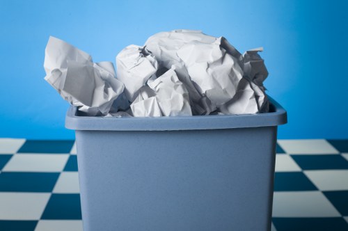 In the image, there is a rectangular light blue plastic bin filled with crumpled white paper on a checkered floor with alternating white and dark blue tiles. The paper is piled high, with some pieces protruding over the top edge of the bin. The background features a plain, bright blue wall, creating a clean and simple environment. The setting appears to be indoors, possibly in a storage or waste collection area. The crumpled paper exhibits varying textures and shades of white and light gray, with some parts appearing more wrinkled and others more smooth. The overall scene suggests a collection of paper waste ready for disposal or removal, consistent with professional rubbish removal services. This image is suitable for illustrating waste collection, rubbish management, or disposal tasks carried out by Commercial Waste Removal Norwood in the south London area, near Norwood or surrounding postcode districts.