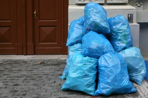 A group of large, light blue plastic rubbish bags filled with waste, piled on a paved pavement in front of a brown wooden door and nearby wall in an outdoor urban setting. The bags appear to be tightly knotted at the top, with some slightly bulging indicating contents inside. The surface around the bags consists of grey stone or concrete pavers, and the background features a smooth, painted wall with a utility box or electrical outlet visible nearby. The scene suggests a commercial waste collection area, with the bags prepared for rubbish removal services, consistent with the offerings of Commercial Waste Removal Norwood near the postcode area of Norwood, London.
