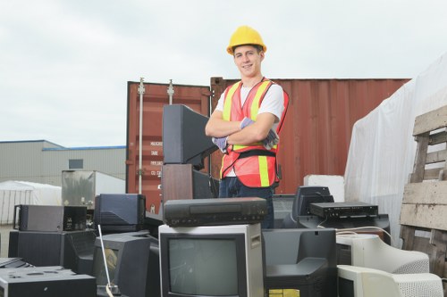 A young male worker wearing a yellow safety helmet and a high-visibility safety vest with orange and yellow stripes stands outdoors in an industrial yard, surrounded by a variety of discarded electronic appliances including old computer monitors, CPUs, and other electronic waste. The worker is smiling and has his arms crossed, facing the camera. Behind him, there are large shipping containers and a stack of wooden pallets near a warehouse building, indicating a waste disposal or recycling site typical of rubbish clearance operations in the Norwood area. The condition of the appliances shows signs of dust and wear, with some devices stacked on top of each other in a somewhat disorganized manner. The environment suggests a professional waste management setting where Commercial Waste Removal Norwood handles electronic and general rubbish collection, contributing to proper disposal practices in the local community. Bright daylight and a cloudy sky provide natural lighting, highlighting the details of the electronic waste and the workerâ€™s safety gear against the industrial backdrop.
