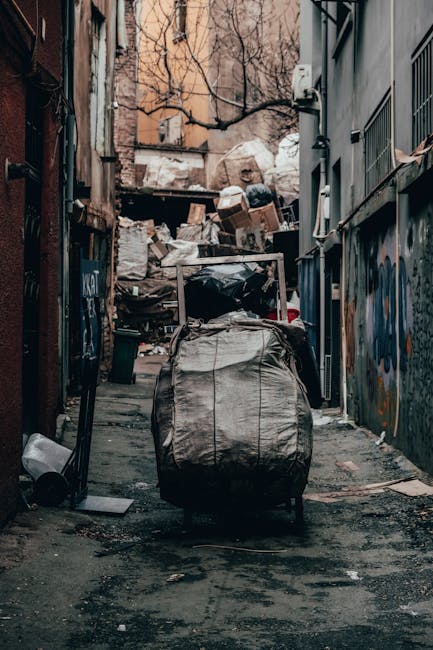 A large, weathered red metal waste container situated on a dark, textured pavement corner between two building walls. To the right of the container, several transparent plastic rubbish bags, filled wi