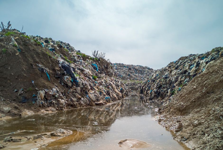 A narrow, muddy water channel runs through a heavily debris-strewn construction or landfill site, with steep, uneven soil embankments on both sides covered in scattered waste materials. The waste cons