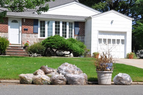A residential street scene featuring a suburban house with a white garage door and a brick and stone facade, situated in Norwood. In the foreground, on the curb, there is an arrangement of large, irregularly shaped rocks with rough textures and light grey and brown tones. Next to the rocks, a grey metal bucket contains dried, brown, withered plants, with some smaller rocks placed beside it. The house has a well-maintained green lawn and a neatly trimmed shrubbery under the front bay window. A set of brick steps leads up to the front door, and a concrete driveway extends to the garage. Tall trees with leafy branches cast shadows across the scene, creating a bright and clear atmosphere typical of a sunny day in the local area. This image visually aligns with services offered by Commercial Waste Removal Norwood, illustrating the type of waste or debris that might be handled in residential rubbish removal efforts.