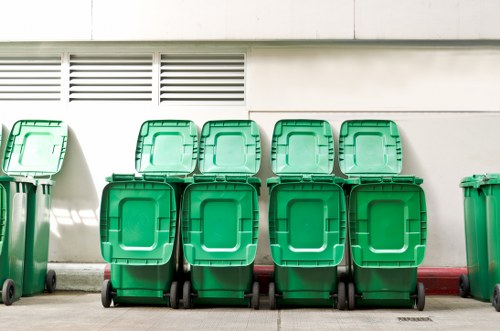 A row of eight green plastic wheelie bins lined up on a paved surface in front of a white building with small, rectangular ventilation slats near the top. The bins have hinged lids with ridged surfaces and are equipped with black wheels at the base. They are positioned closely together, with some bins slightly overlapping others, and are likely used for waste collection or rubbish disposal services in the local area. The scene appears to be outdoors, possibly on a driveway or service yard, with natural daylight illuminating the bins and casting subtle shadows along the wall behind them. The overall setting suggests an organized waste management area typical for commercial or residential rubbish collection services, such as those provided by Commercial Waste Removal Norwood, serving the local postcode area. The bins' vibrant green colour contrasts with the neutral background, emphasizing their function within the rubbish removal context.